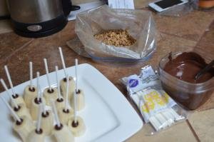 A plate with banana halves with lollipop sticks in them, a plastic bag with crushed walnuts, a bag of lollipop sticks and a bowl of melted chocolate on a table