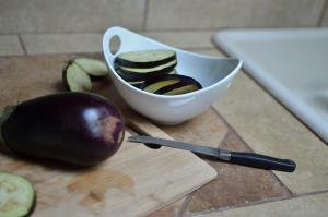 Full eggplant on a cutting board with horizontally cut circle slices in a bowl next to it