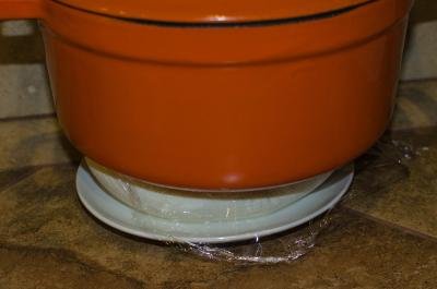 Farmer's Cheese on a plate wrapped in plastic wrap with a heavy ceramic pot standing on top of it