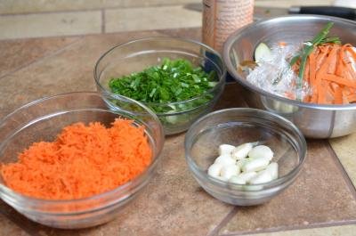 Ingredients on table include a bowl of grated carrots, sliced scallions, and peeled garlic