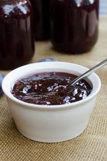 Prune Plum Jam in a bowl with a spoon and prunes around it, with jars behind the bowl