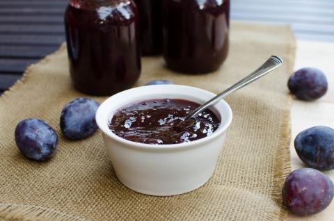 Prune Plum Jam in a bowl with a spoon and prunes around it, with jars behind the bowl