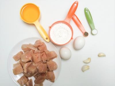 Ingredients on table include; chicken cut into bite size cubes on a plate, 2 eggs, 3 garlic cloves, slat and pepper, cornstarch and oil