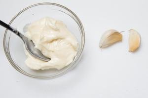 A bowl with mayo and with 2 garlic cloves next to the bowl
