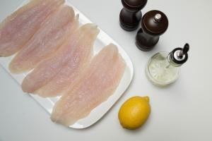 On the table there is a plate with 4 fish fillets, a lemon, oil, and slat and pepper