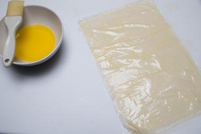 A bowl of melted butter with a pastry brush in it and a sheet of filo dough laid out next to the bowl