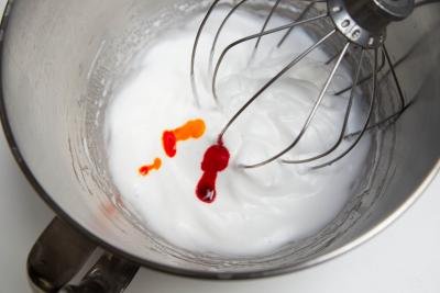 Egg whites and sugar being beat in a KitchenAid mixer with red and yellow food coloring added it the mixture