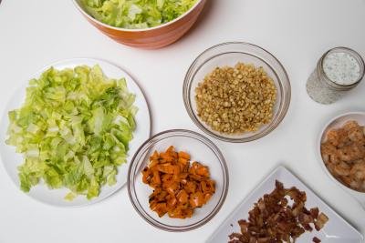 Ingredients on table including; chopped up lettuce on a plate, roasted peppers in a bowl, roasted corn in a bowl, fried bacon on a plate, shrimp in a bowl and each dressing in a jar