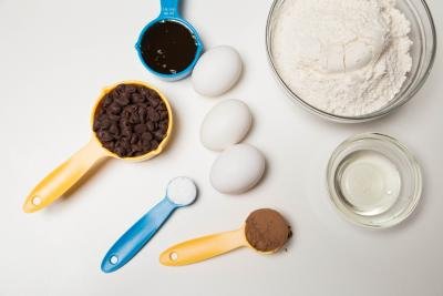 Ingredients on table including; 3 eggs, a bowl with flour, a bowl with Irish liqueur, a measuring spoon with chocolate chips, another with cocoa, the third with baking soda , and fourth with honey