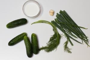 Ingredients on table including 4 cucumbers, dill, green onion, 3 cloves of garlic, and a bowl with sour cream