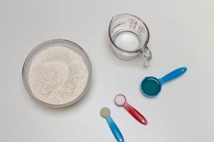 Ingredients on table including; a large bowl with flour, a measuring cup with water, and 3 measuring spoons one with oil, another with yeast and third with salt