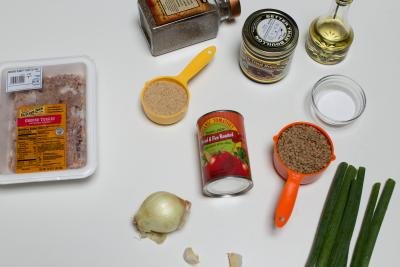 Ingredients on the table including; an onion, green onions, 2 garlic cloves, a can of tomatoes, a measuring cup with lentils, a small bowl salt, a measuring cup with bread crumbs, a jar of better than boullion, oil, pepper, and ground turkey