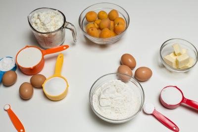 Ingredients on the table including; flour in a bowl, 6 eggs, a bowl with butter, a bowl with apricots, a measuring cup with farmers cheese, and 6 measuring spoons with sugar, baking powder, whipping cream, potato starch