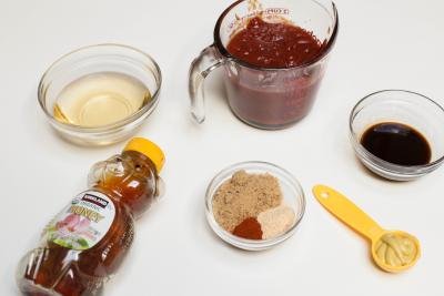 Ingredients on table including; honey, a little bowl with sider vinegar, a bowl with Worcestershire sauce, a bowl with brown sugar, paprika and garlic powder, a measuring cup with ketchup and a measuring spoon with honey mustard