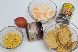 Ingredients on table including; corn in a bowl, chips in a bowl, spicy clack bean dip in a jar, a tomato, a jar of pinto beans, and a bowl of cheese
