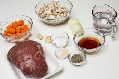 Ingredients on table including; beef, carrots in a bowl, mushrooms in a bowl, a bowl with marsala wine, 2 full onions, a measuring cup of water, garlic, salt and pepper and garlic salt in little bowls