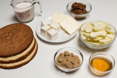 Ingredients on table including; cake slices, a bowl of brown sugar, a bowl of apple juice, a bowl of peeled and sliced apples, a bowl with dulce de leche, a plate with butter and cream cheese and a measuring cup with heavy whipping cream