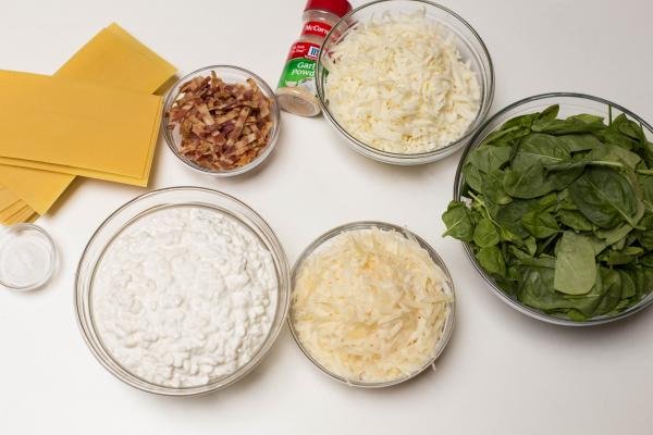Ingredients on table including; a bowl with cottage cheese, a bowl with mozzarella, another bowl with parmesan, a bowl with spinach, a little bowl with bacon, garlic powder, a bowl with salt and dry lasagna pasta