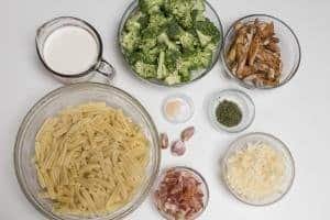 Ingredients on the table including; 5 bowls one with pasta, another with cheese, one with chicken, third with broccoli and fifth with bacon, a measuring cup with heavy whipping cream, and a little bowl with salt and garlic powder