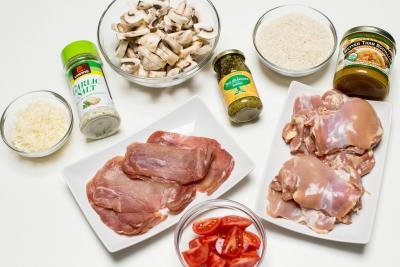 Ingredients on the table including; a plate with chicken thighs, a plate with prosciutto, a bowl with sliced tomatoes, a bowl with sliced mushrooms, a bowl with rice, a bowl with cheese, a jar of garlic salt, a jar of basil pesto, and a jar of better than bouillon chicken
