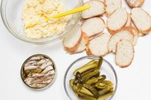 Table with sliced baguette and pickles, a bowl with egg and mayo mixture, and sprats