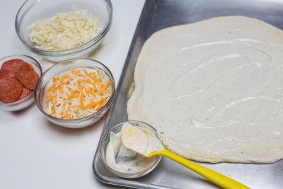Ranch being spread on rolled out dough that is laying on the baking pan, with 2 bowls besides the pan, one with cheese other with pepperoni \
