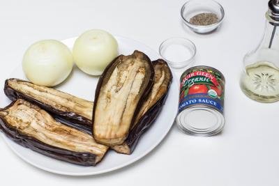 Ingredients on the table including; 2 halved eggplants on a plate, a jar of tomato sauce, 2 onions, a little bowl of salt and one with pepper