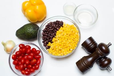 Ingredients on the table including; an onion, avocado, a yellow bell pepper, a bowl with baby tomatoes, a bowl with black beans and corn, salt and pepper, and 2 little bowls one with lemon juice and one with oil