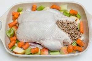 Seasoned duck in a ceramic baking pan filled with buckwheat with apples, carrots and celery around it