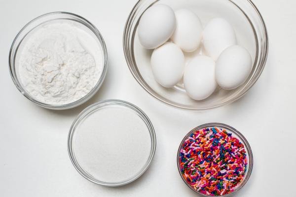 4 bowls on the table on with sprinkles, one with sugar, one with eggs and last one with flour