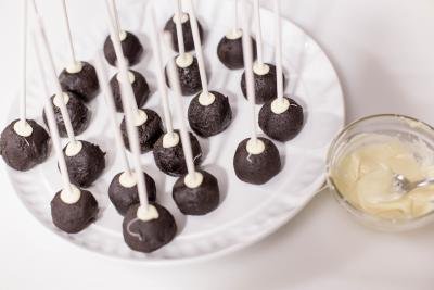 Cake pop stick being dipped into a bowl with white chocolate and then into the oreo balls on a plate