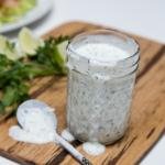 Light Ranch Dressing in a jar on a cutting board with herbs and lime slices behind the jar and a spoon in front of the jar