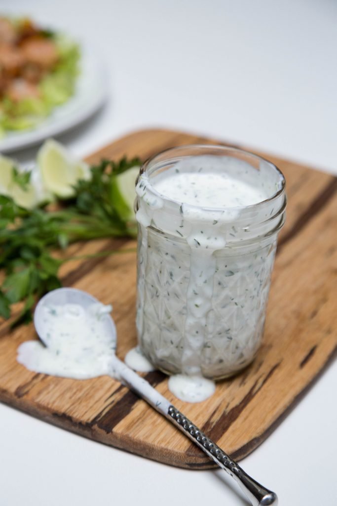 Light Ranch Dressing in a jar on a cutting board with herbs and lime slices behind the jar and a spoon in front of the jar