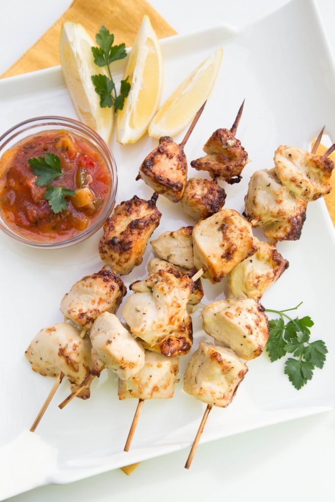 Chicken Kebabs on a plate with lemon slices next to it and salsa in a small bowl next to the kebabs