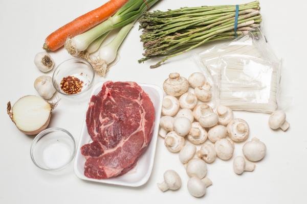 Ingredients on the table including; mushrooms, rice noodles, beef, half an onion, a bowl of salt, a bowl of red pepper, garlic, green onions, a carrot and asparagus