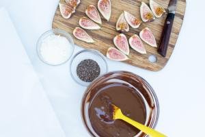 On the table a cutting board with figs, a bowl with chocolate, a bowl with chia seeds and a bowl with coconut flakes