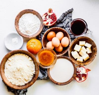 7 bowls on the table with the following ingredients; almond flour, sugar, raw honey, butter, eggs, baking powder, and all purpose flour also on the table an orange, 2 pomegranate halves, and a measuring cup with pomegranate juice