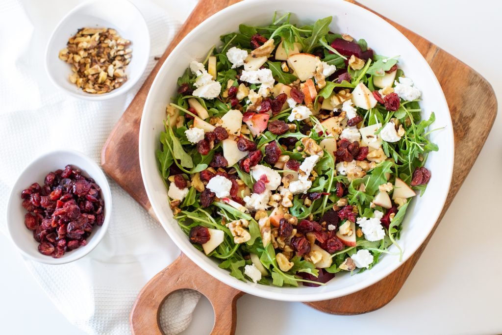 Beet and Goat Cheese Arugula Salad in a bowl with a bowl of dried cranberries and a bowl of walnuts besides it