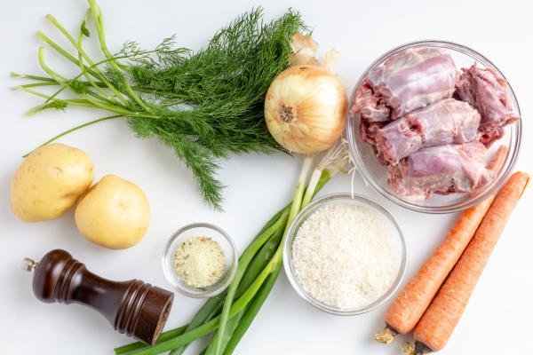 Ingredients on the table including; a bowl of turkey necks, 2 carrots, green onions, a bowl of rice, a bowl of seasoning, pepper, 2 potatoes, an onion and dill