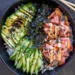Rice in a bowl with salmon, avocado, seaweed, cucumbers and sesame seeds with sauce.