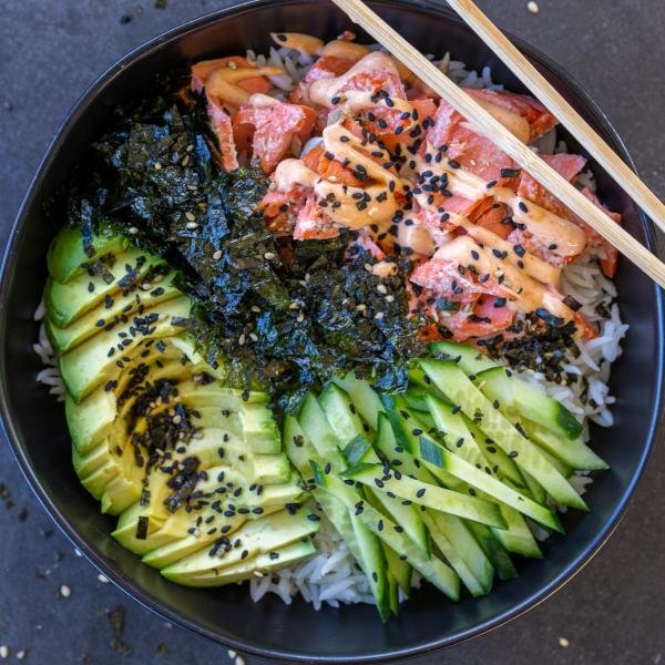 Rice in a bowl with salmon, avocado, seaweed, cucumbers and sesame seeds with chop sticks.