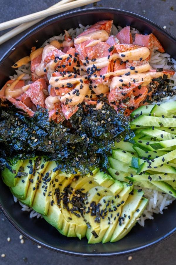 Rice in a bowl with salmon, avocado, seaweed, cucumbers and sesame seeds adn sauce. 