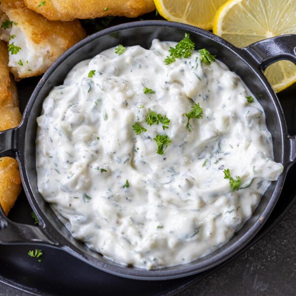 Tartar sauce in a serving dish with herbs and lemon next to it.