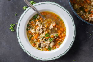 A bowl of Chicken Barley Soup with spoon and herbs around.