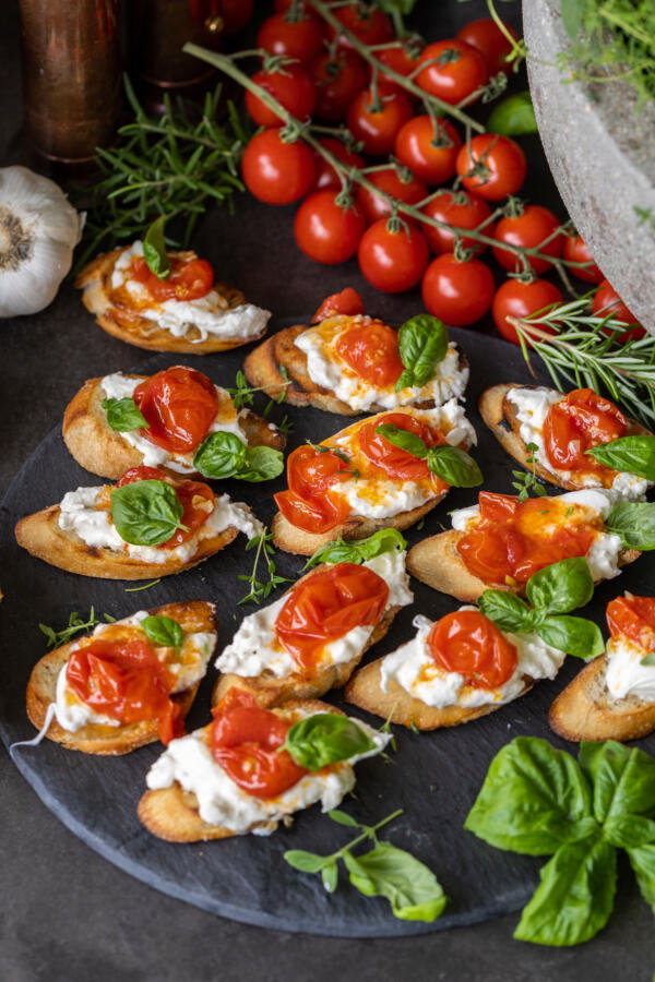 Burrata Crostini with Blistered Tomatoes on a tray and other ingredients from recipe around. 